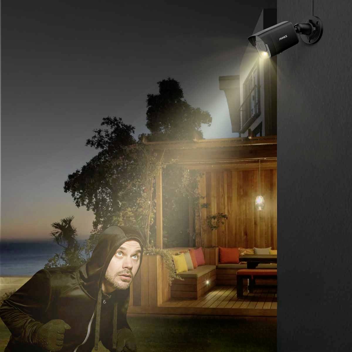 A man in dark clothing approaches a lit house at night, while a security camera with light monitors him.