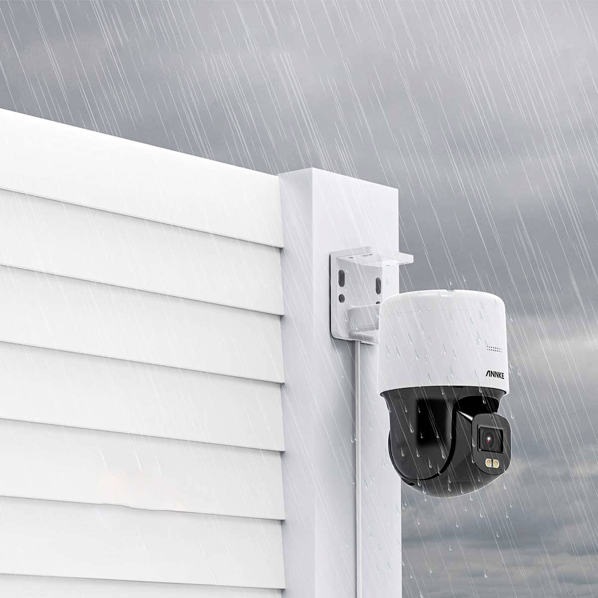 A surveillance camera mounted on a white wall in the rain. The camera is weatherproof and aimed at the exterior area.
