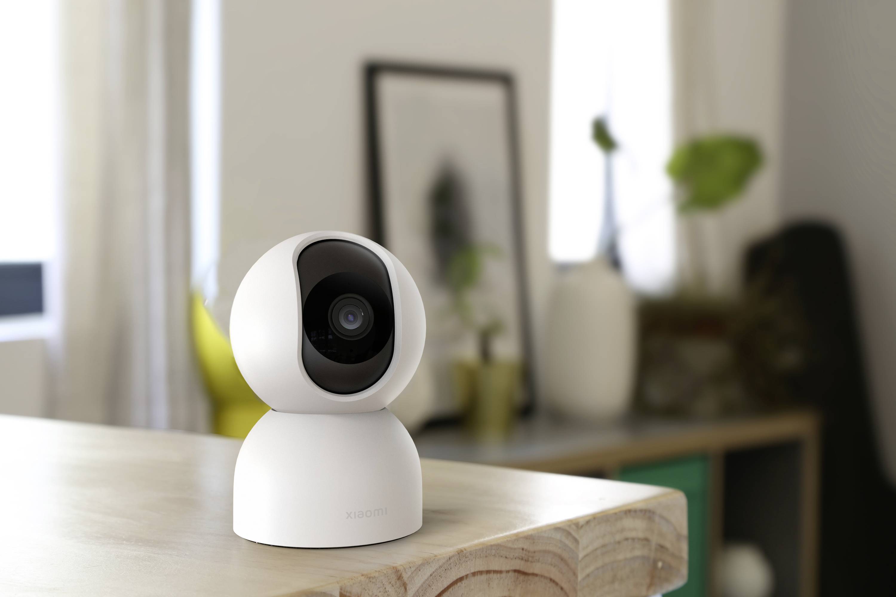 A white surveillance camera sits on a table in a light-filled room with a blurred background and plants.