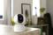 A white surveillance camera sits on a table in a light-filled room with a blurred background and plants.