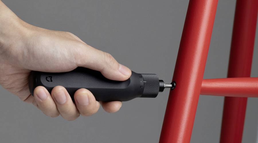 A hand is holding a black, electric screwdriver and tightening a screw in a red piece of furniture.