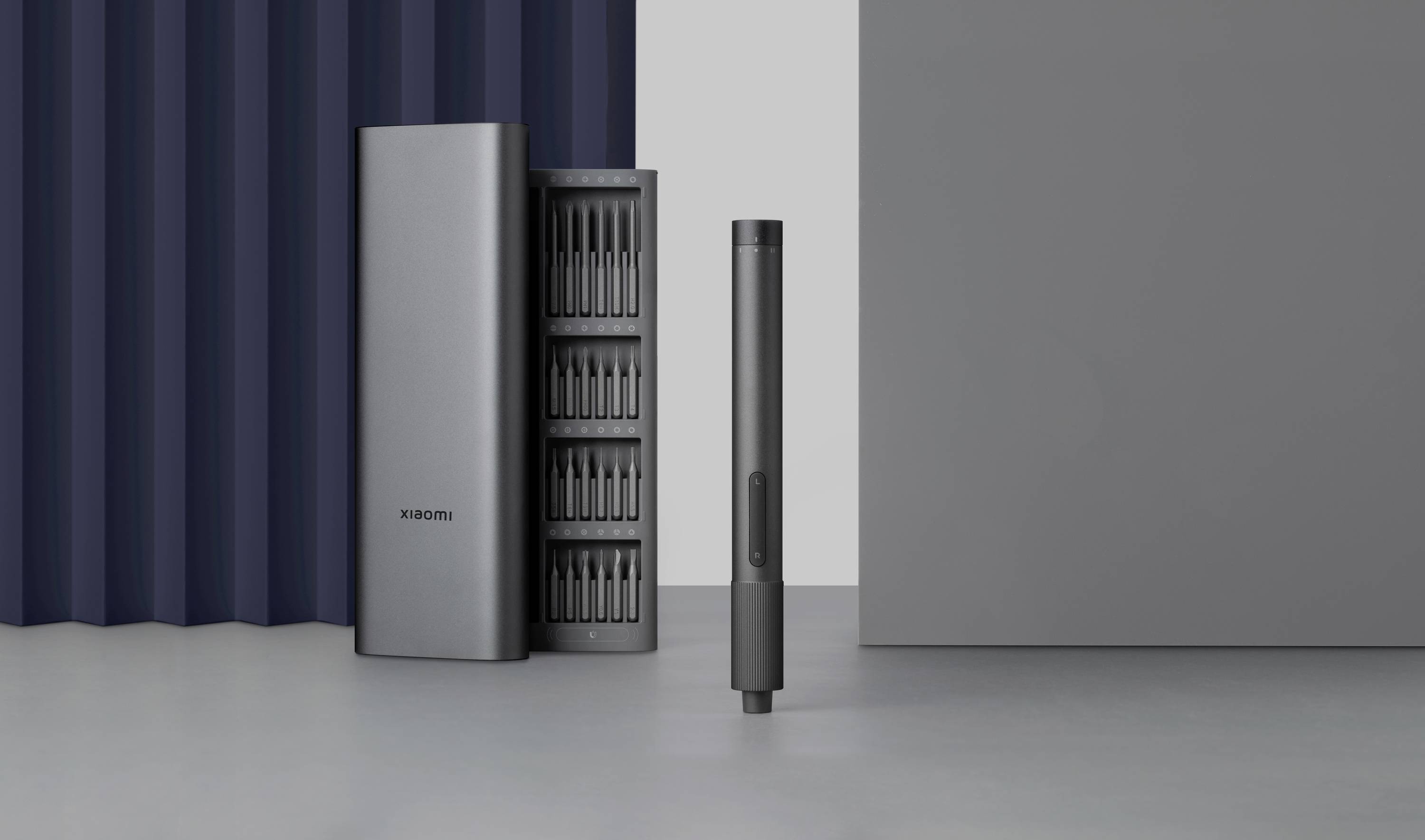 A dark grey, slender screwdriver with a stand base. In the background, a matching open case with multiple inserts.