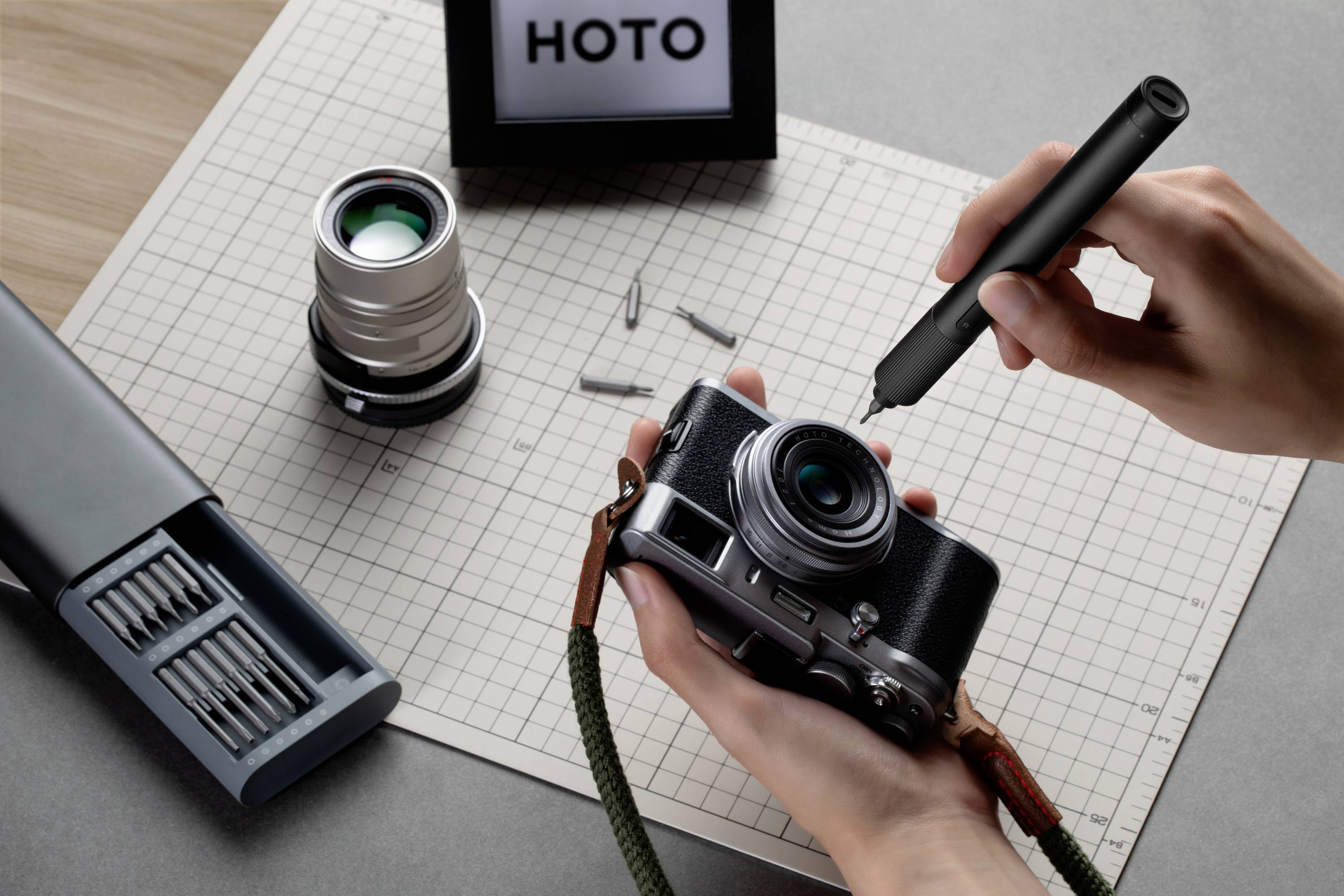 A person is repairing a camera with a screwdriver on a desk. Two camera lenses and screws are lying beside it.