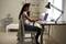 A woman is sitting at a desk in a bright room, working on a laptop. A bed can be seen in the background.