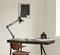 A simply designed workspace with a dark wooden table, featuring a black desk lamp, books, and a small jug. An abstract artwork hangs on the wall in the background.