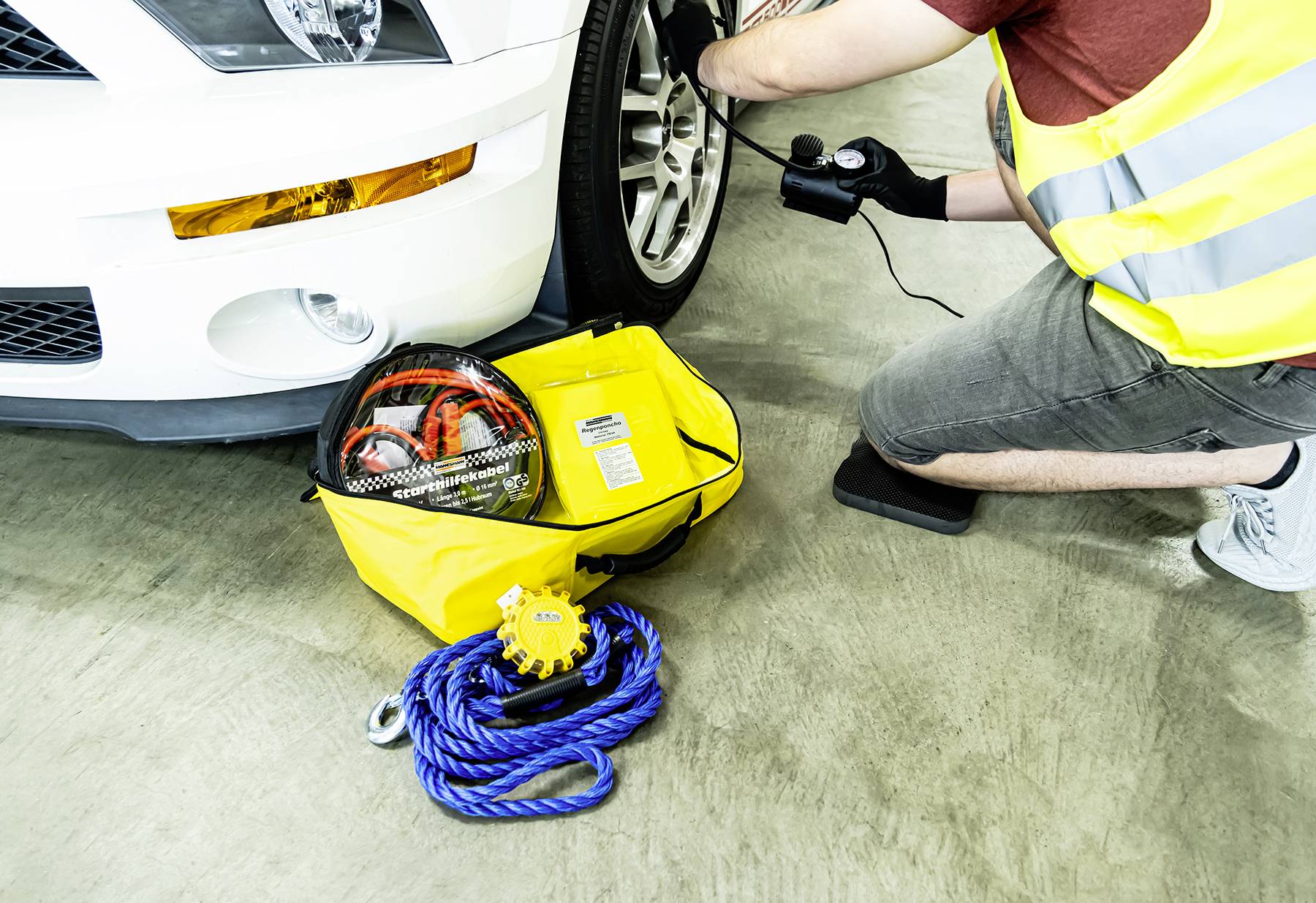 A person wearing a high-visibility vest is checking the tyre pressure of a white car. In the foreground, a yellow bag with emergency equipment is lying down.