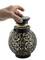 A hand removes the lid of a decorative round lantern with an intricate filigree pattern. The lantern is black and has a glass insert.