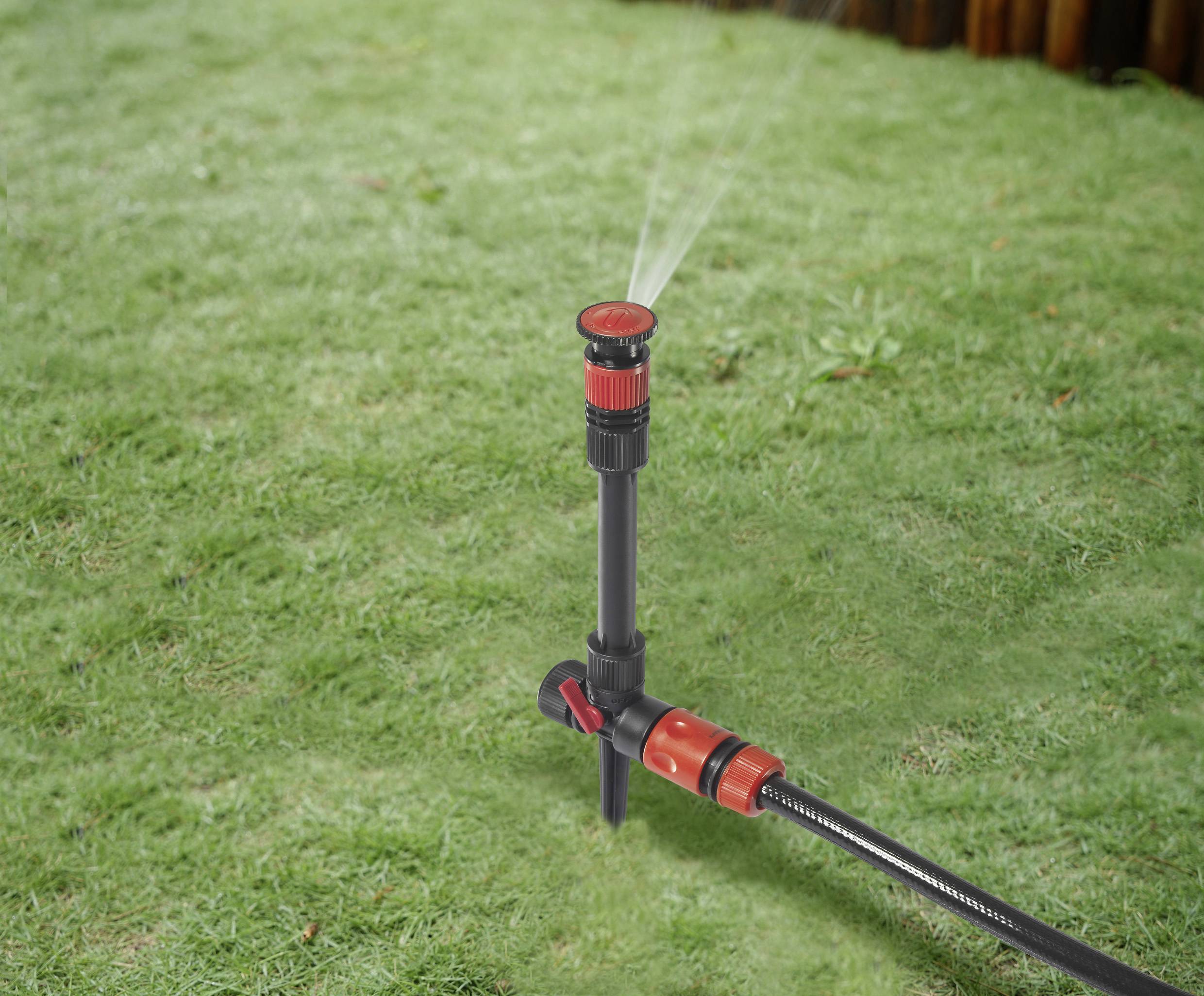 A lawn sprinkler waters a green lawn evenly. The sprinkler head is black and red and connected to a hosepipe.