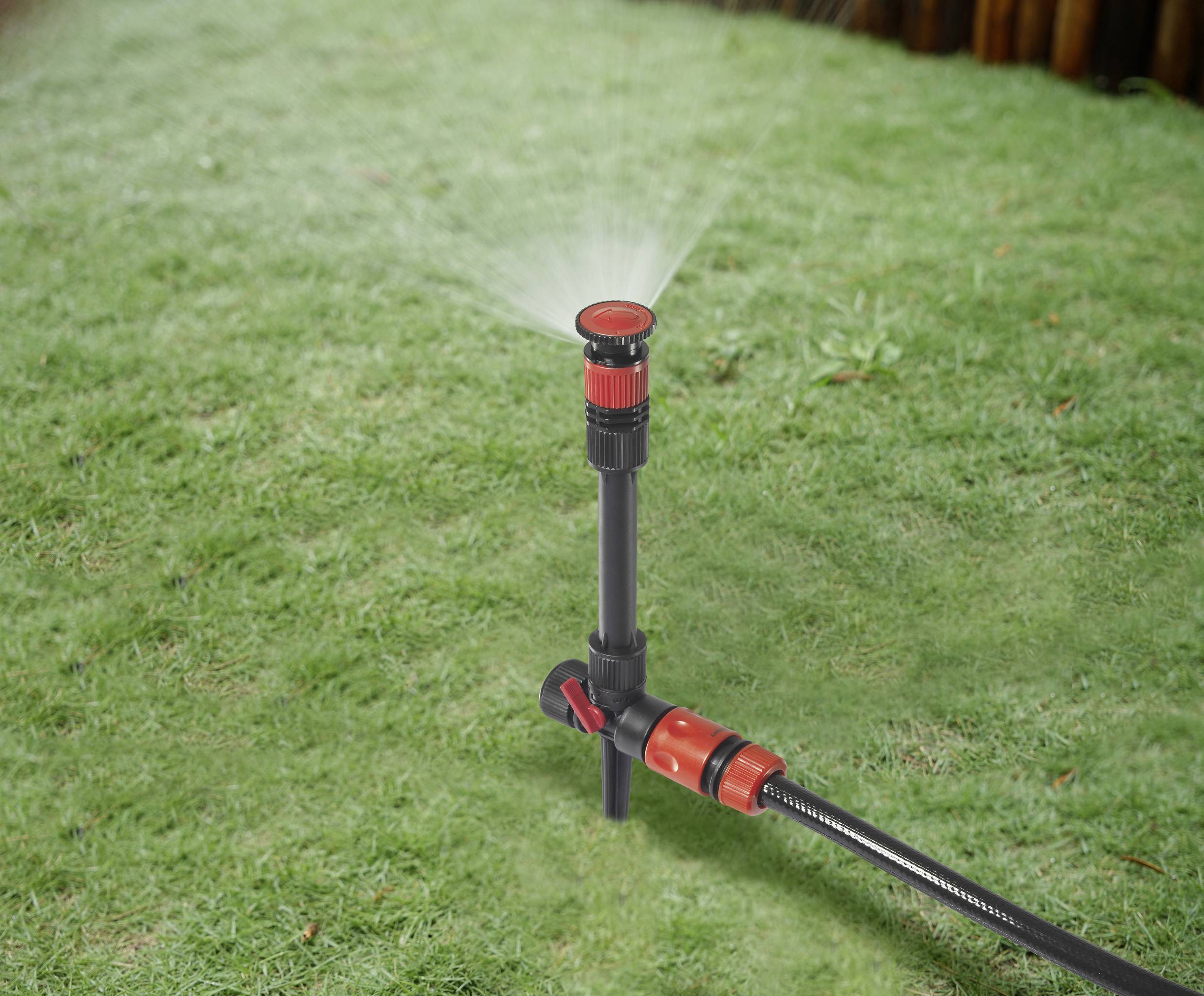 A lawn sprinkler on a grassy area distributes water evenly. The sprinkler head is red and black, connected to a hosepipe.