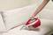 A person is cleaning a beige sofa with a portable, red vacuum cleaner.