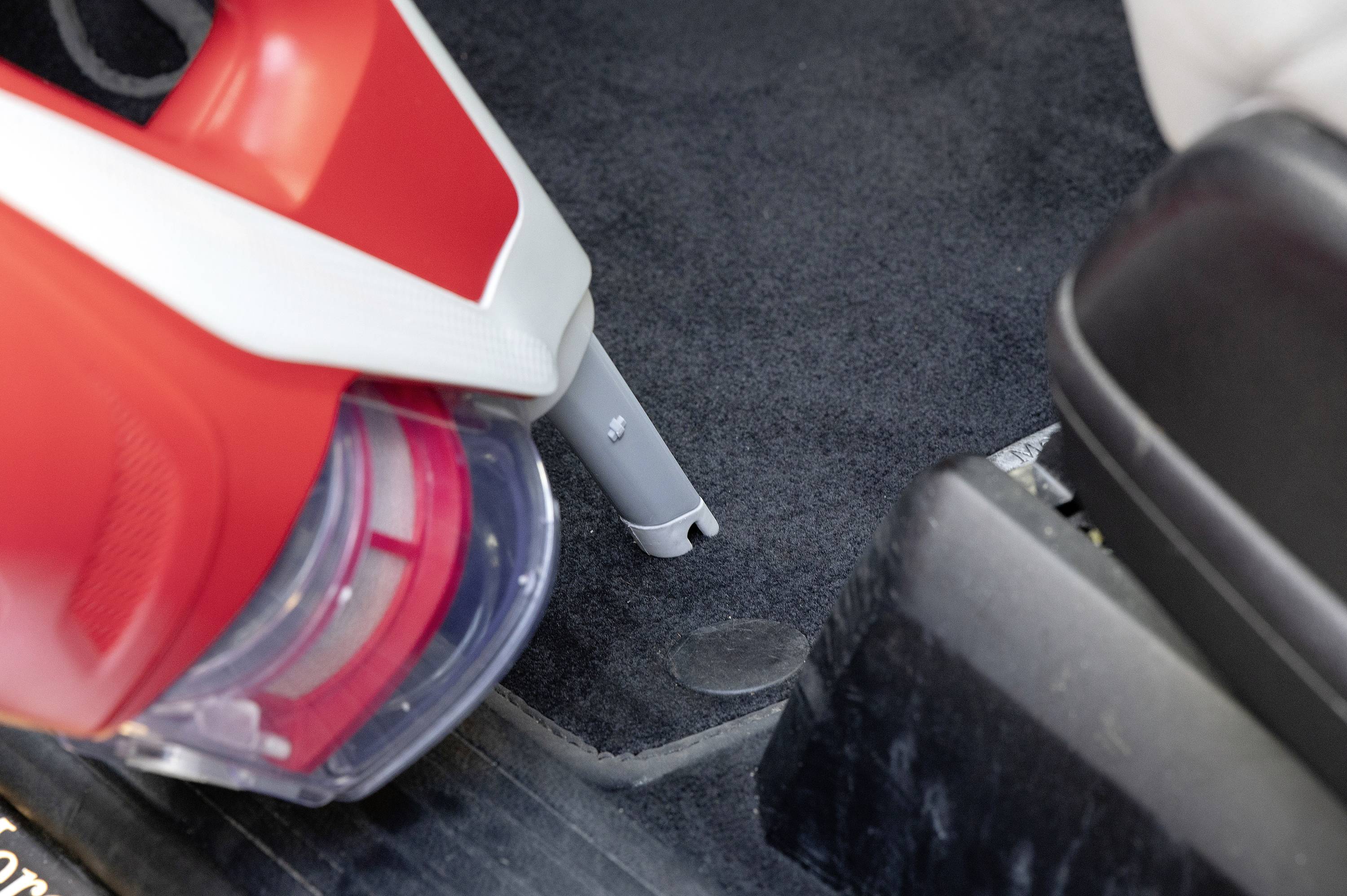 Vacuum cleaner with a red casing hoovering a car carpet beside the seat; focus on the narrow nozzle and the suction movement on the floor.