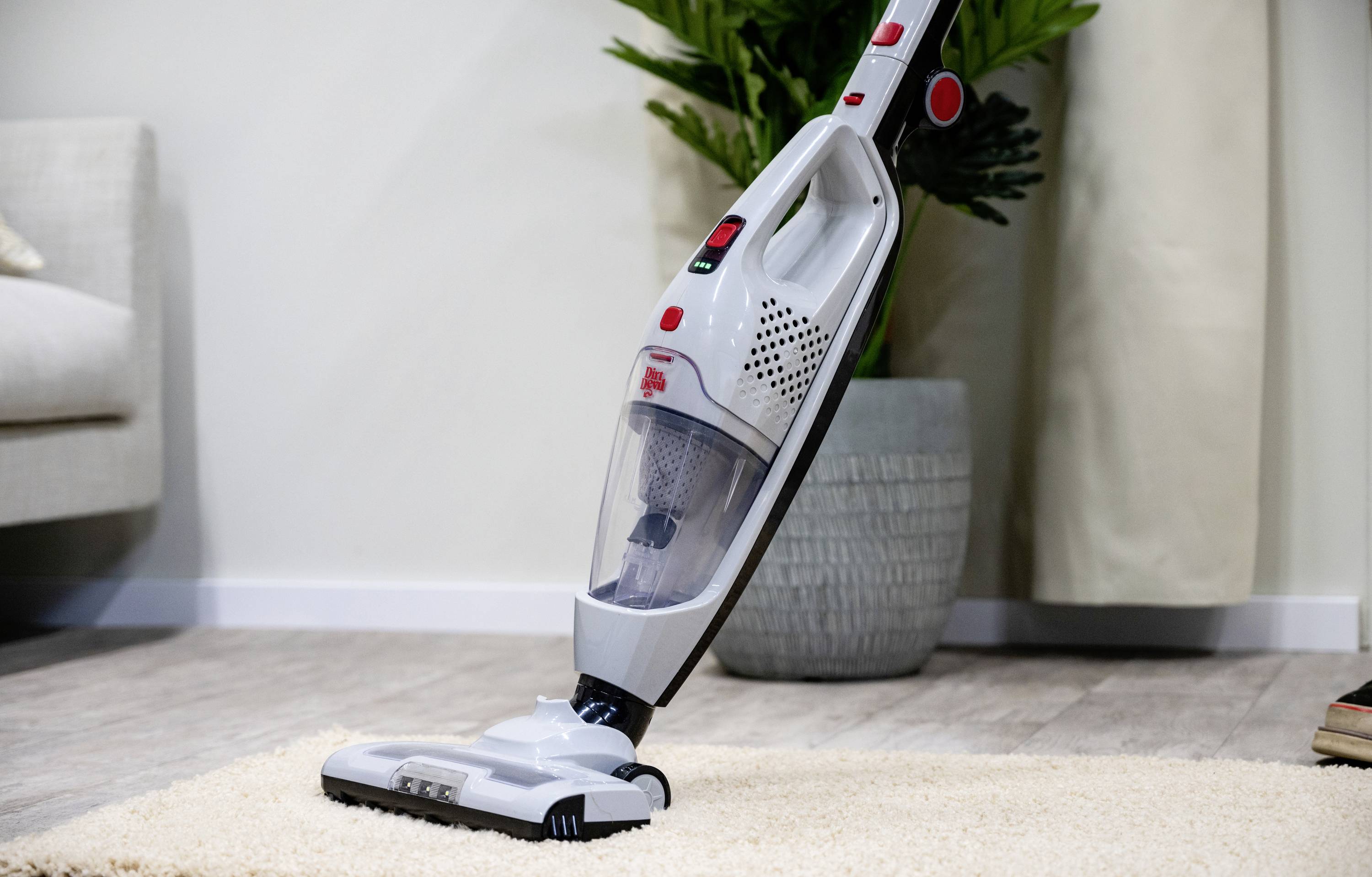 A white vacuum cleaner is cleaning a beige carpet in a living room with a wooden floor and a plant in the background.