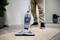 A person is using a vacuum cleaner on a beige carpet in a room with wooden flooring and plants in the background.