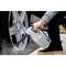 A person is holding a steam cleaner, cleaning a car wheel rim. They are wearing jeans and shoes with a thick sole. The tyre is in focus.
