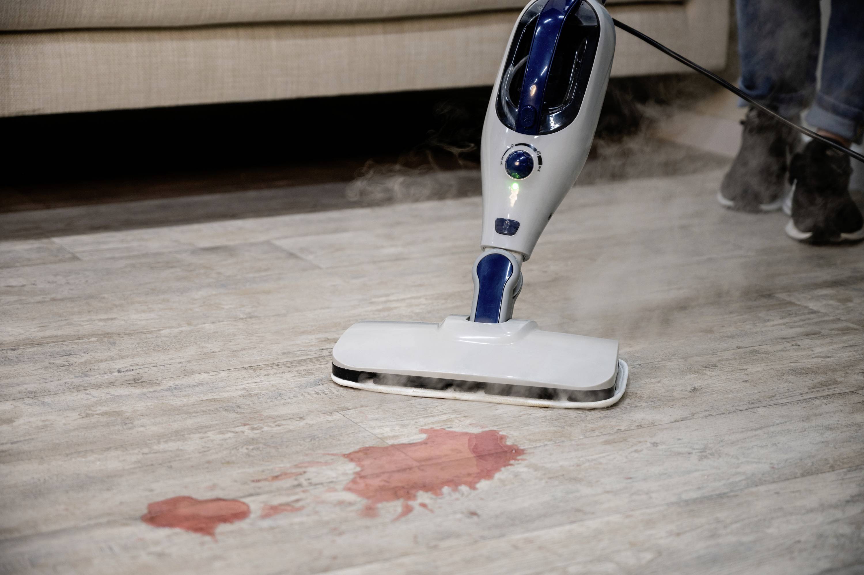 Steam cleaner removes red stain from wooden floor. Sofa and person blurred in the background. Focus on cleaning process.