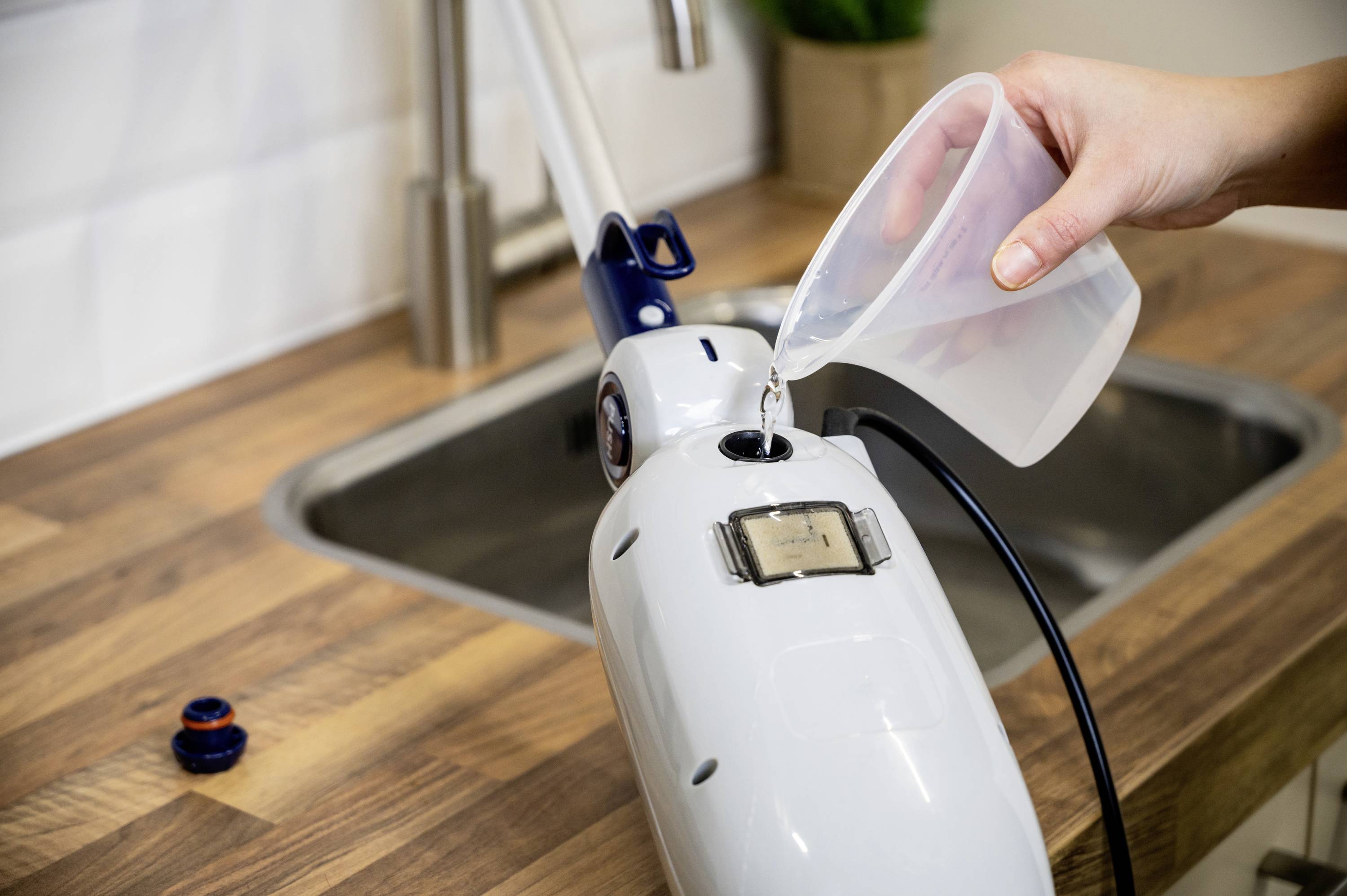 A person is filling water into a steam cleaner container. The open cap is resting on a kitchen worktop next to a sink.