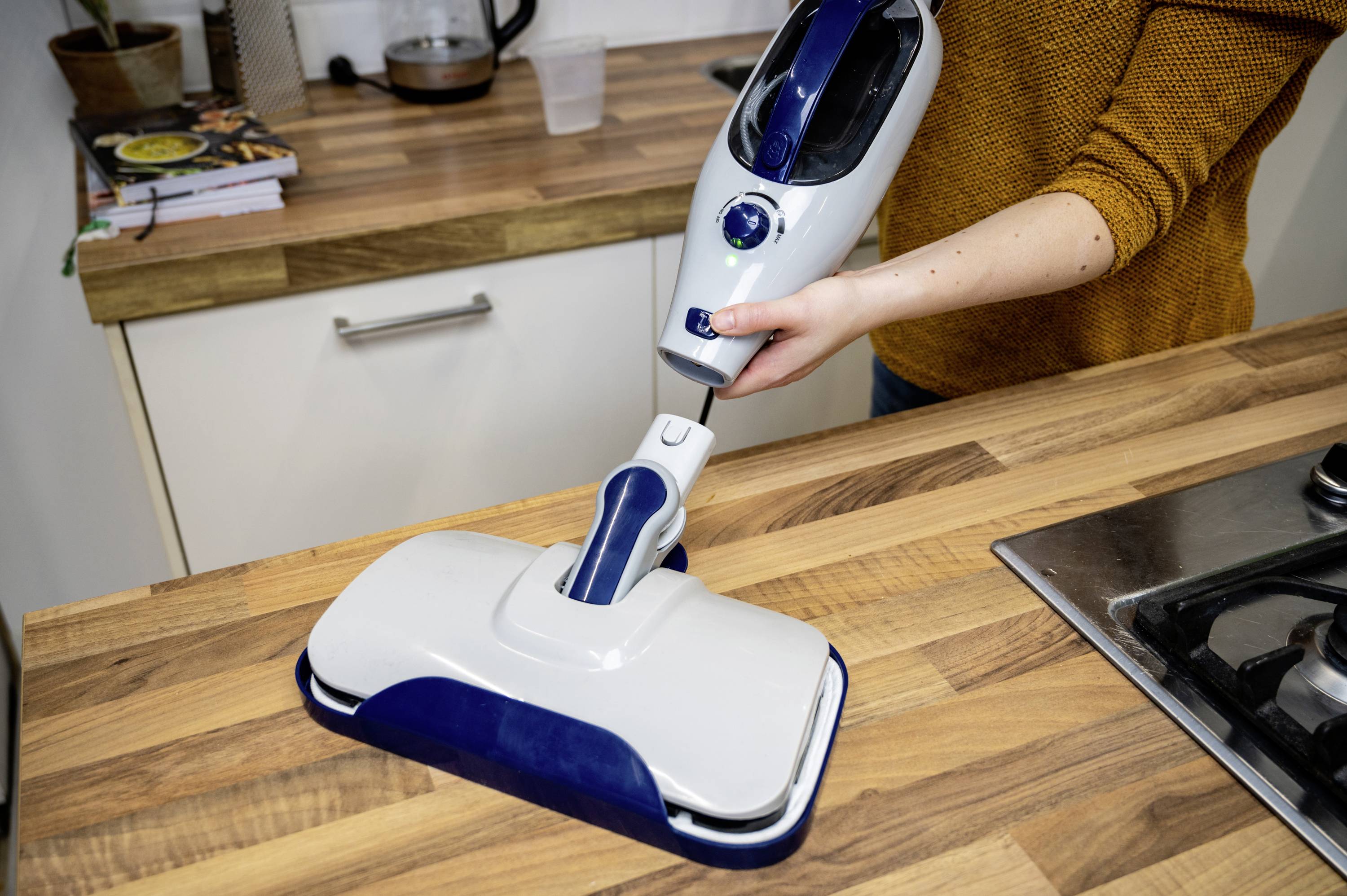 A person is using a steam cleaner on a wooden worktop in a kitchen. Books and kitchen appliances can be seen in the background.