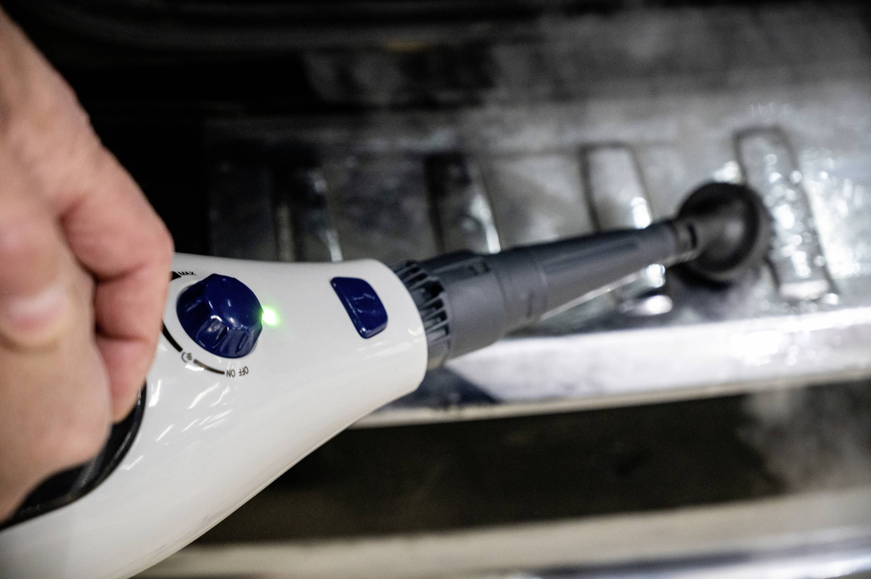 A steam cleaner nozzle is cleaning a metal surface. A hand is holding the cleaner, and the green indicator light is illuminated.