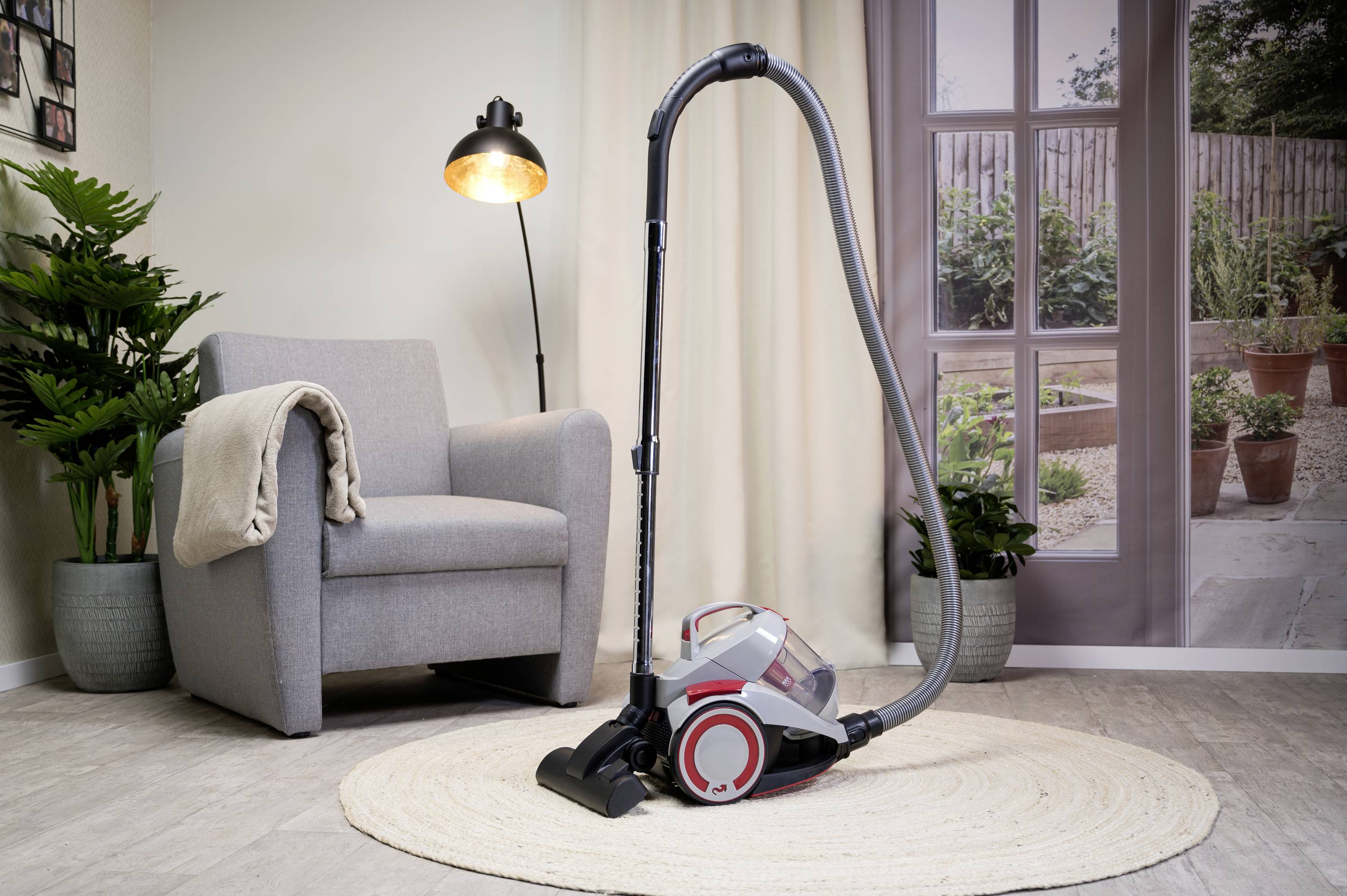 Vacuum cleaner sits on a round rug in a cosy living room with an armchair, lamp, and large windows overlooking the terrace.