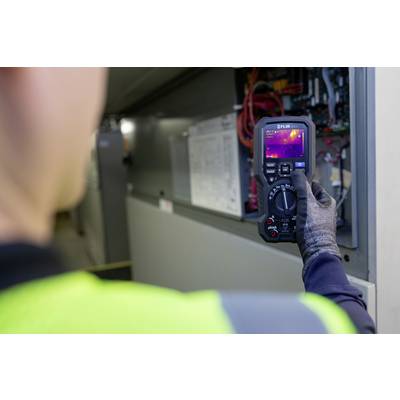 A technician in protective clothing is inspecting the electrical components of a control cabinet using a thermal imaging camera.