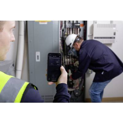 A worker stands in front of an open electrical cabinet and uses a smartphone to check power consumption. A technician is working in the background.