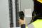 A person is measuring the temperature of a switchboard enclosure using an infrared thermometer. The display shows '27.1°'.