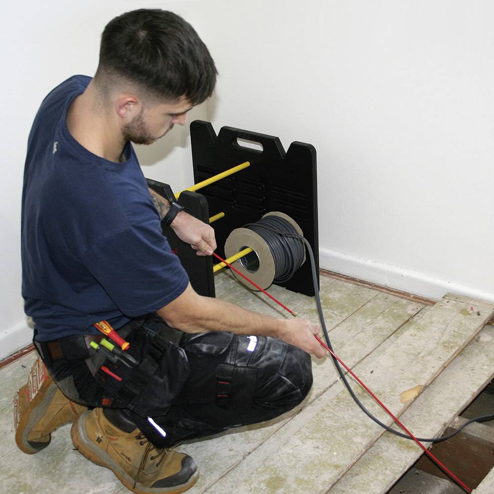 A worker is laying cables in a room. He is kneeling on a wooden floor where some of the floorboards are missing. Next to him lies a cable drum.