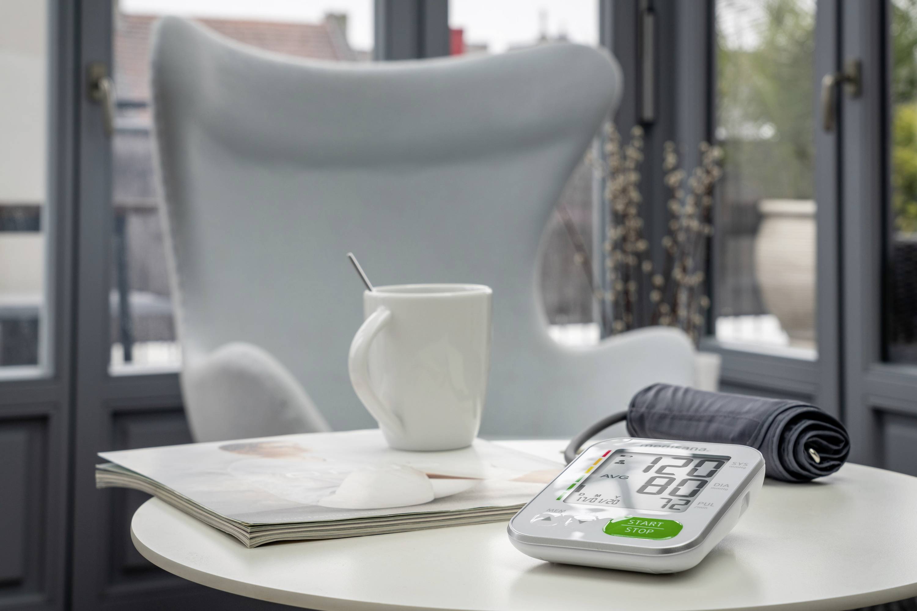 An electronic blood pressure monitor displays 120/80, next to a white cup with a spoon and a magazine on a table.