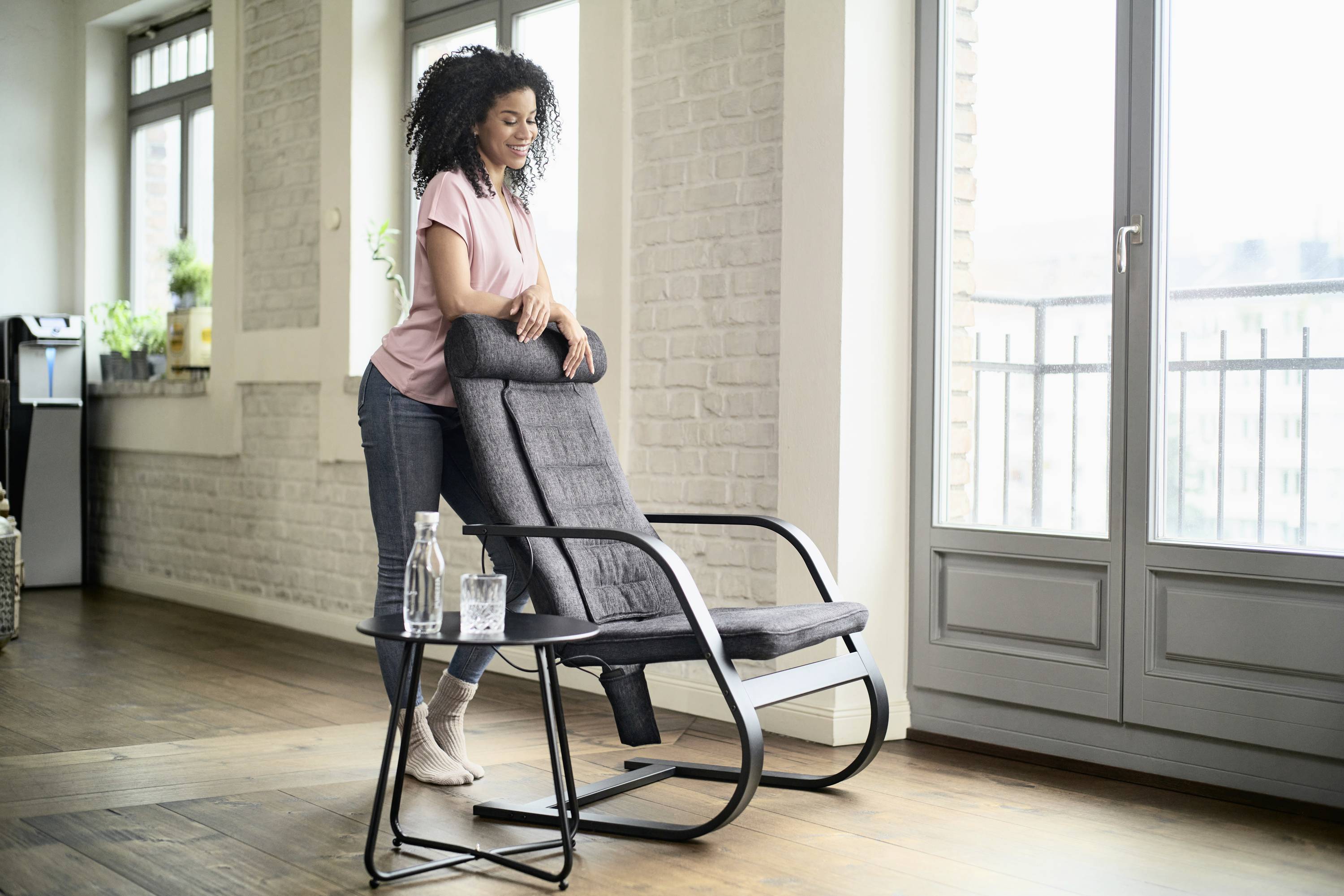 A woman leans against a grey armchair in a bright living room with a wooden floor, beside which stands a table with a carafe and a glass of water.