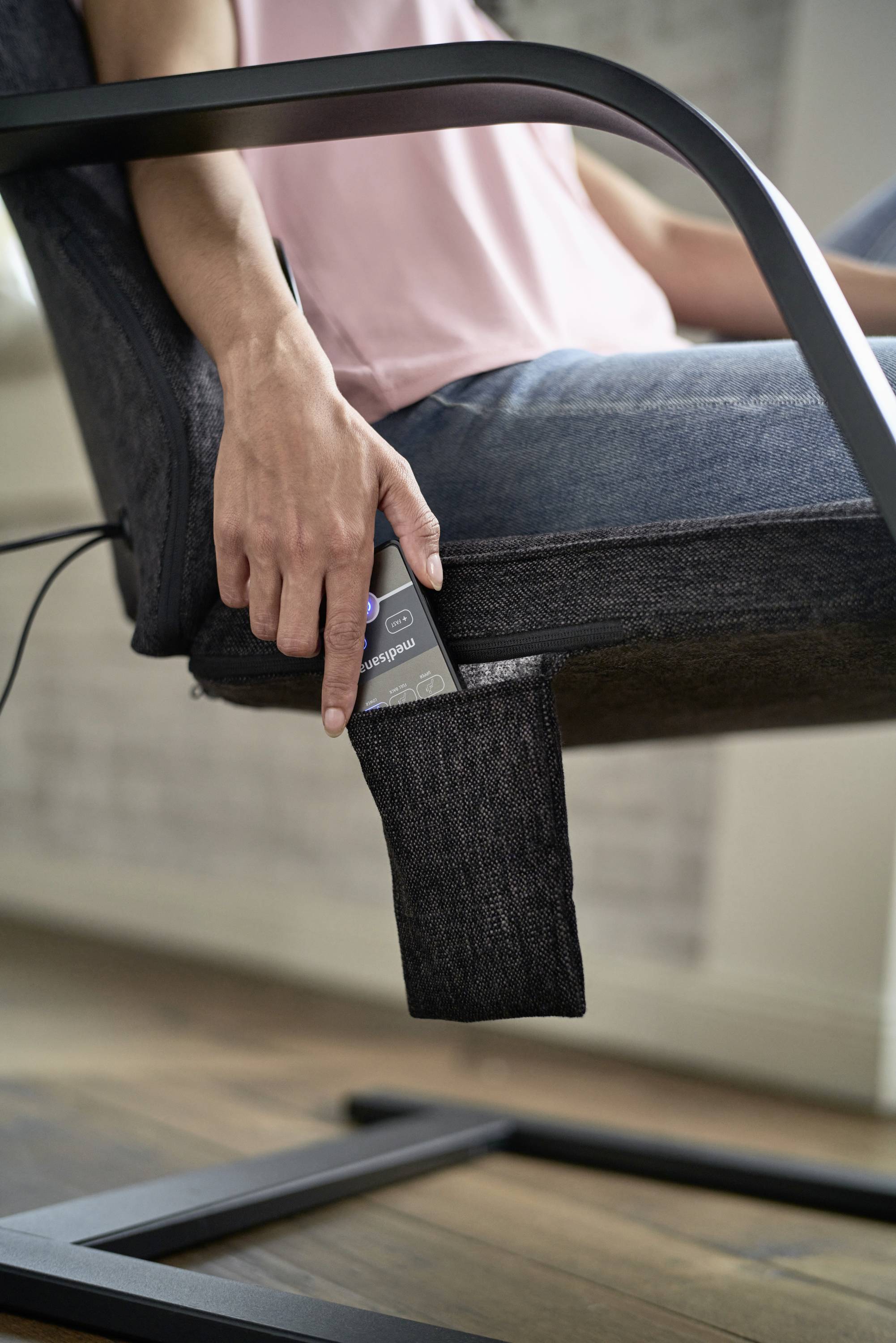 A person reaches for a remote control that is tucked in a fabric bag on the side of a chair.