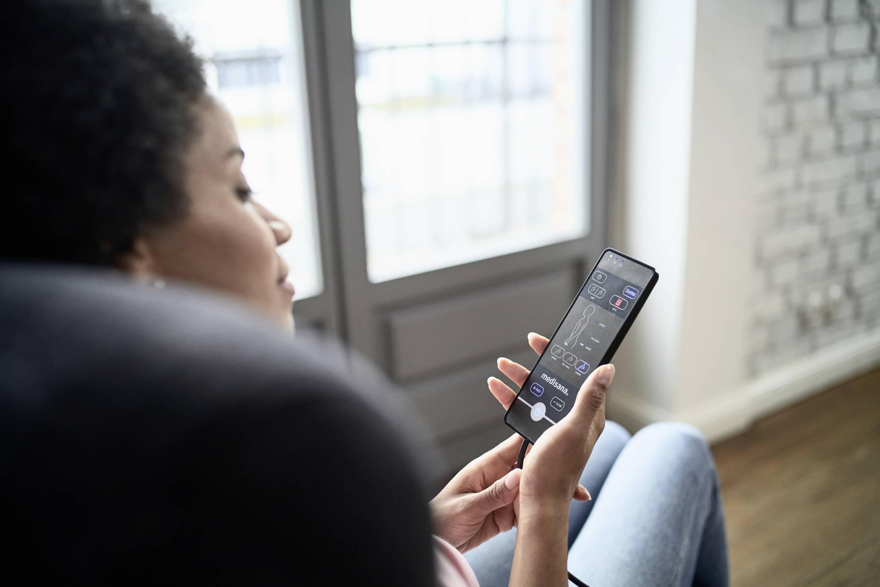 A person is sitting, holding a smartphone in their hand and controlling a smart home system through it. A window is visible in the background.