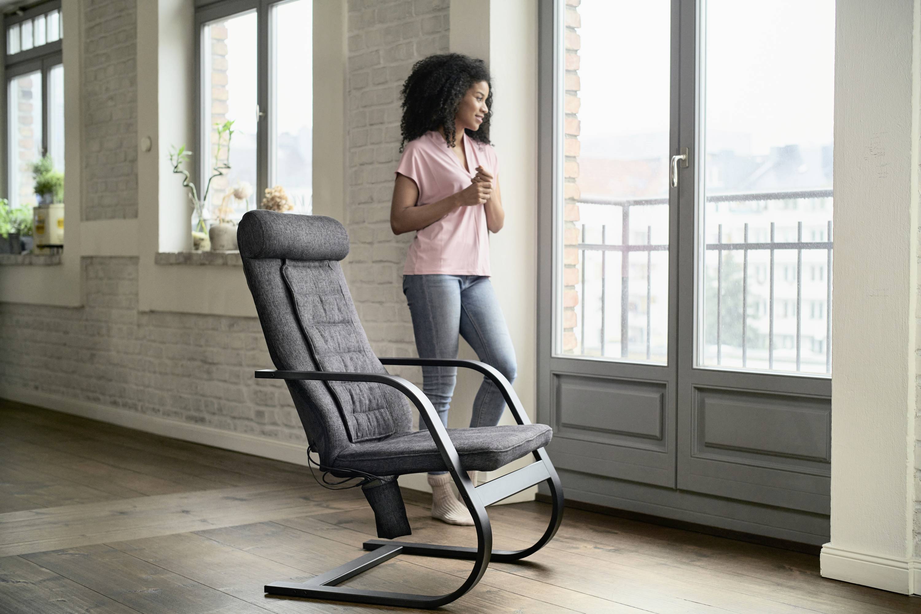 A woman stands in a room in front of a window overlooking an urban landscape. Beside her is an empty, dark grey rocking chair.