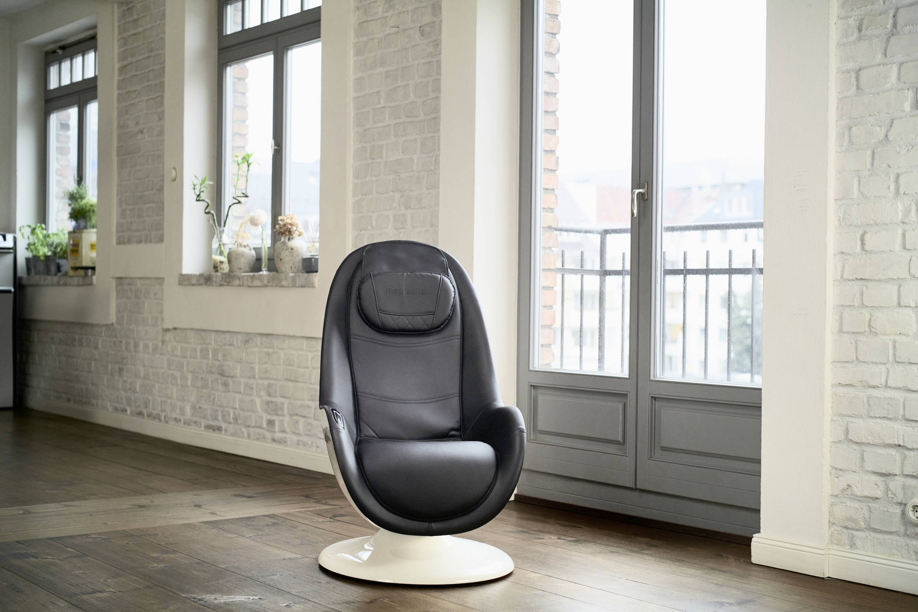 A black, modern armchair sits in a room with pale brick walls and large windows offering a view to the outside.