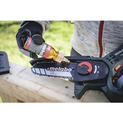 A person is refuelling a chainsaw with oil. The chainsaw is lying on a wooden table. The 'Metabo' brand logo is visible on the saw blade.