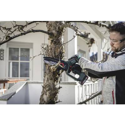A person wearing safety glasses and gloves is cutting branches of a tree with an electric pruning saw in front of a house.