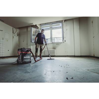 A person is cleaning with a vacuum cleaner in an empty room with bare walls and windows in the background.