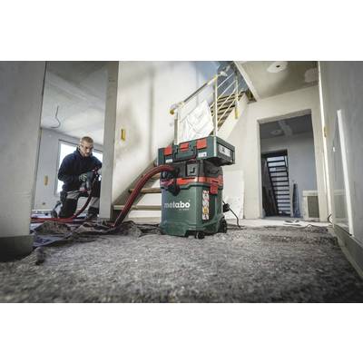 A tradesman is cleaning a construction site with an industrial vacuum cleaner. In the background, a staircase can be seen in an unfinished room.
