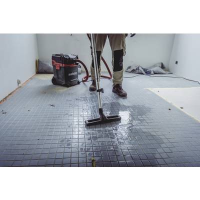 A person is cleaning a wet tiled floor with a vacuum cleaner in an unfinished room. Construction materials are visible in the background.