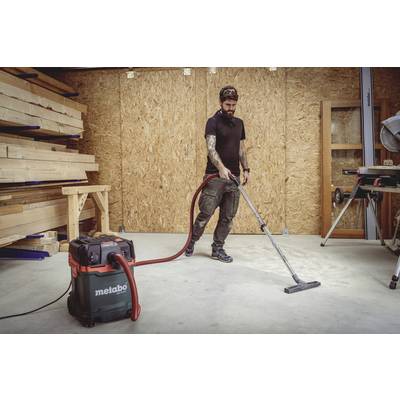 A man is cleaning the floor with an industrial vacuum cleaner in a woodworking workshop. Wooden boards can be seen in the background.