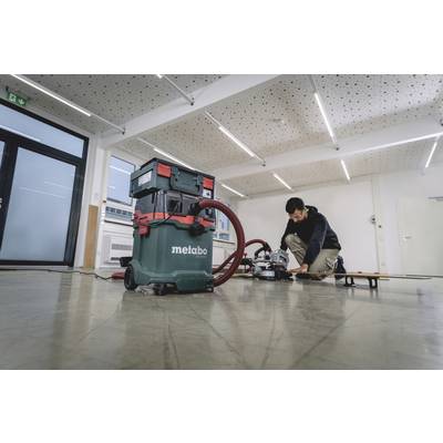 A person is working in a room with a Metabo industrial vacuum cleaner in the foreground. The room has a suspended ceiling with LED lighting.