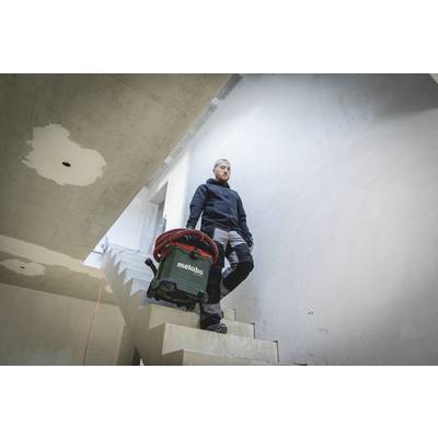 A tradesman is carrying a vacuum cleaner down a staircase in an unfinished building. The surroundings show bare, unplastered walls.