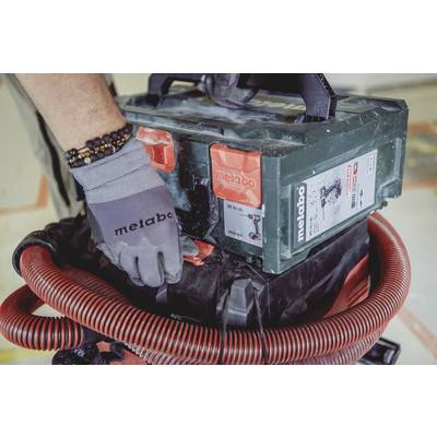 A tradesman is wearing gloves and operating a Metabo device resembling a vacuum cleaner. A red hose is connected to the device.