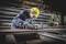 A worker wearing a hard hat and safety glasses is grinding a piece of steel in a metalworking workshop. Metal shelving is visible in the background.