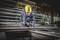 A construction worker in protective clothing and a yellow hard hat is drilling with a machine in a metalworks. Metal scaffolding is visible in the background.