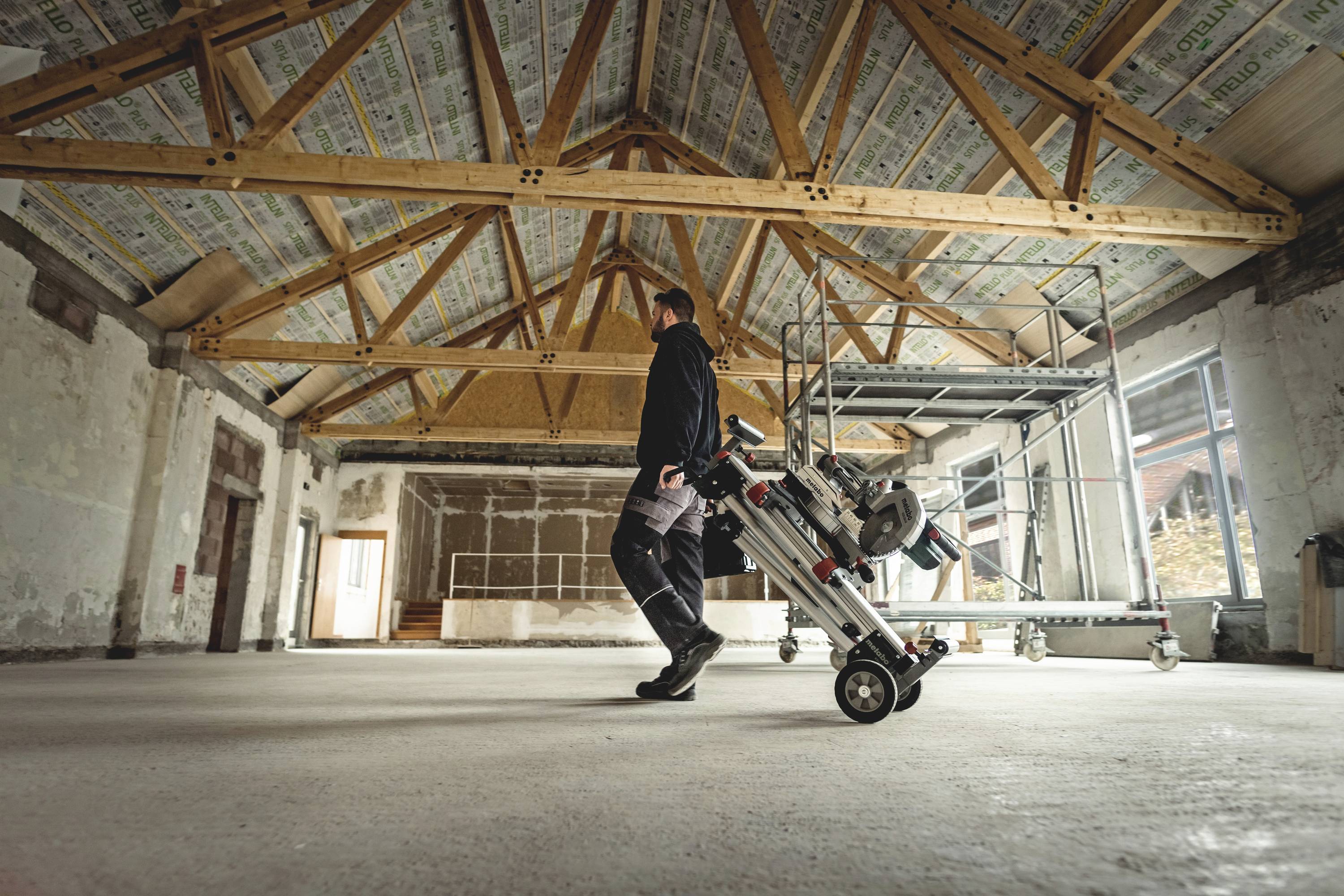 A person in work attire is pulling a wheeled tool through an empty room with an exposed wooden beam ceiling.