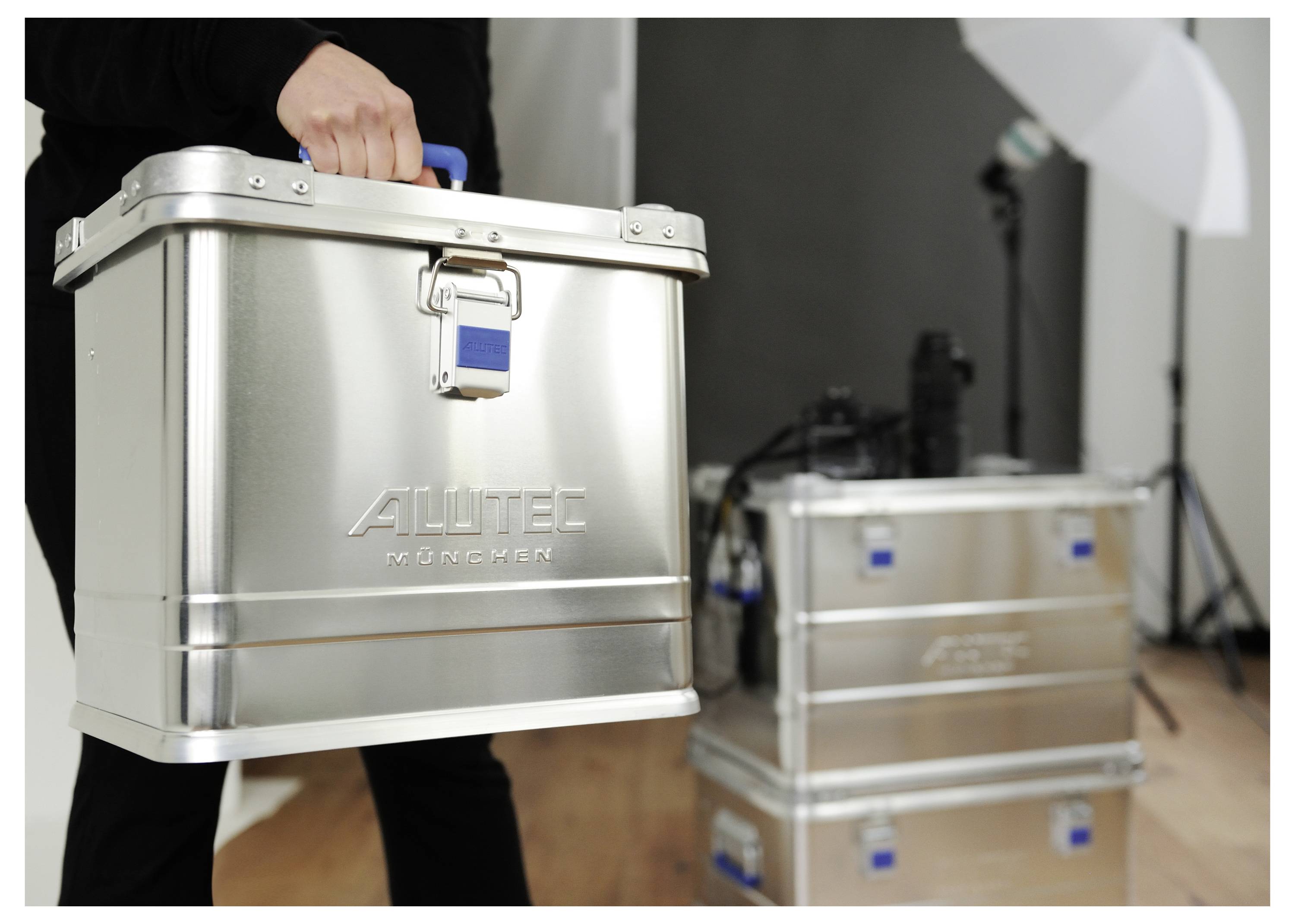 A person carries a silver container with the label 'Aquatec'. Another similar container is in the background. The setting appears to be a studio.