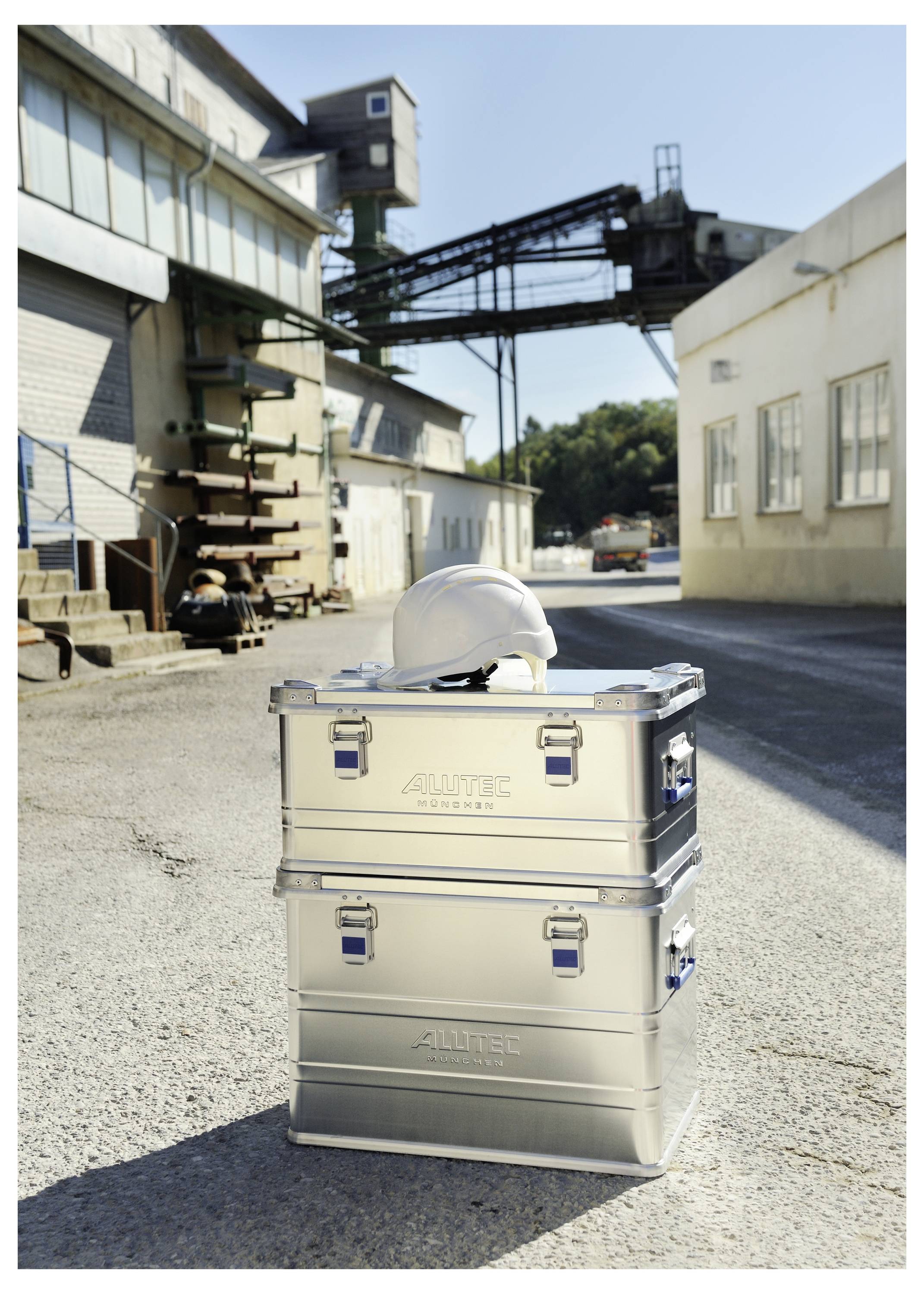 A white hard hat rests on two stacked metal boxes labeled 'Alutec' in an industrial outdoor setting with buildings and machinery in the background.