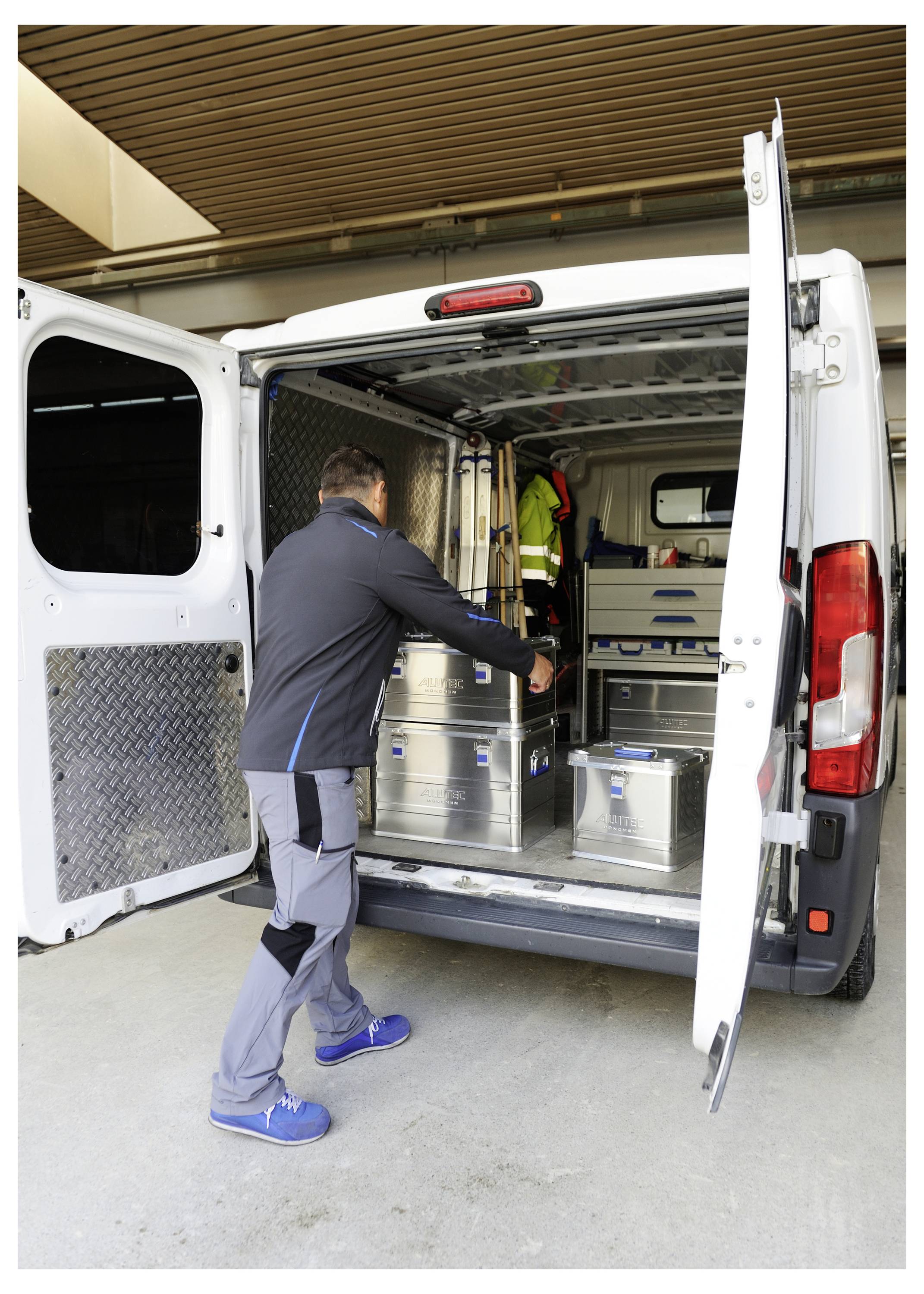 'A person in work attire loads metal containers into a white delivery van parked indoors. The van's rear doors are open, revealing an organized interior.'