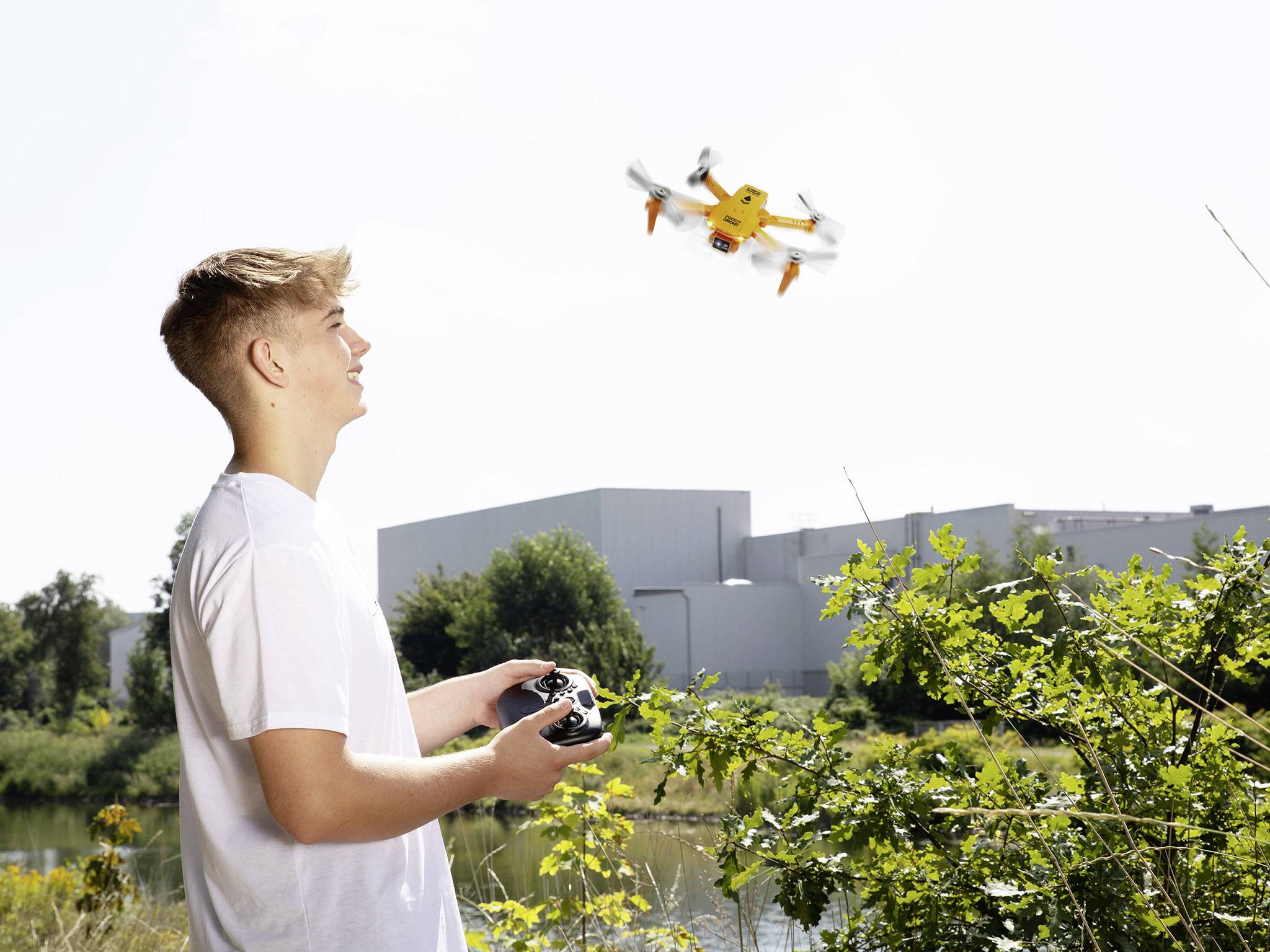 A young man is controlling a flying drone with a remote control beside a river, with trees and buildings visible in the background.