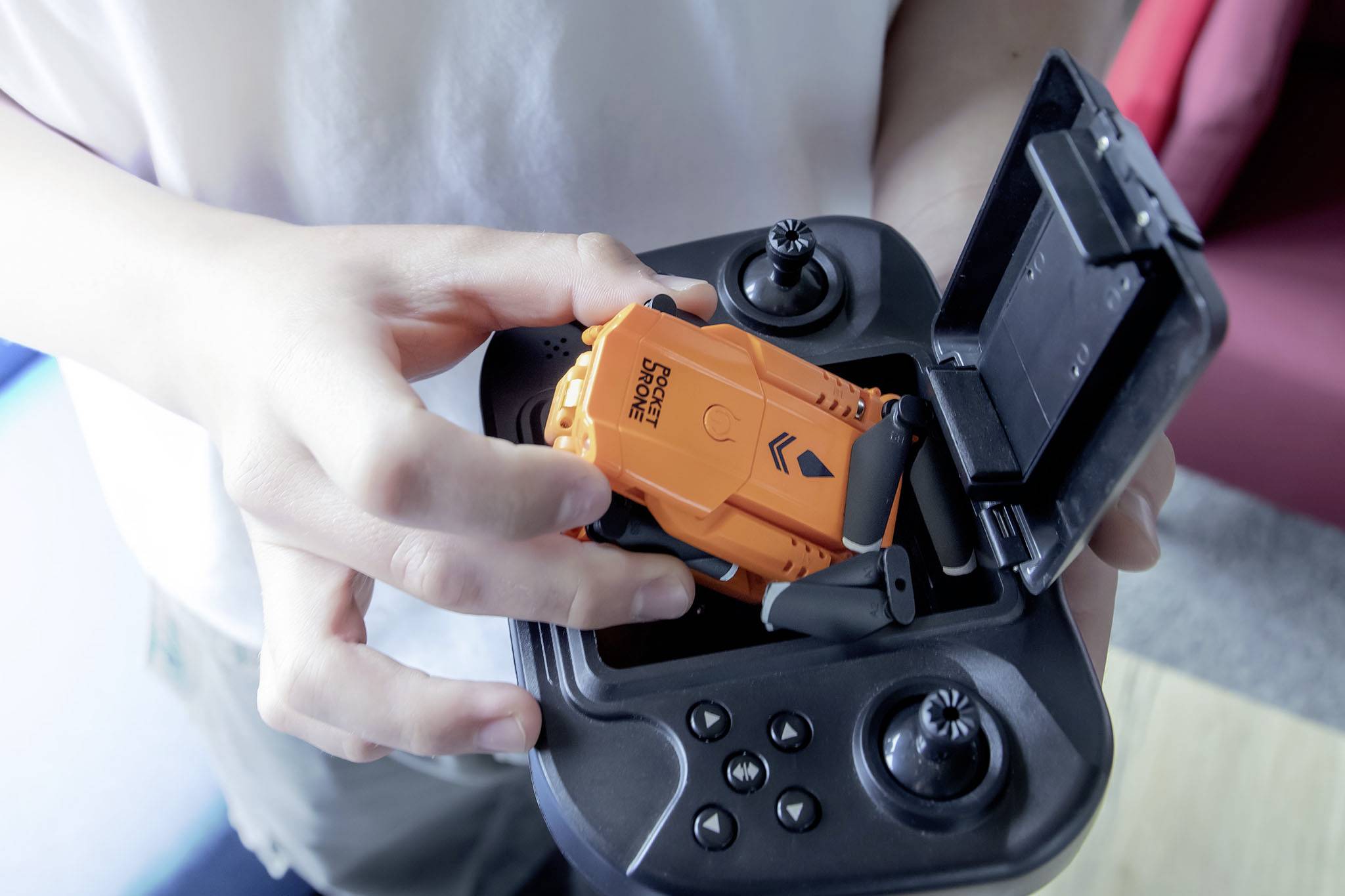 A person is placing a small orange object, possibly a drone with 'DRONE RACING' inscription, into a remote control.
